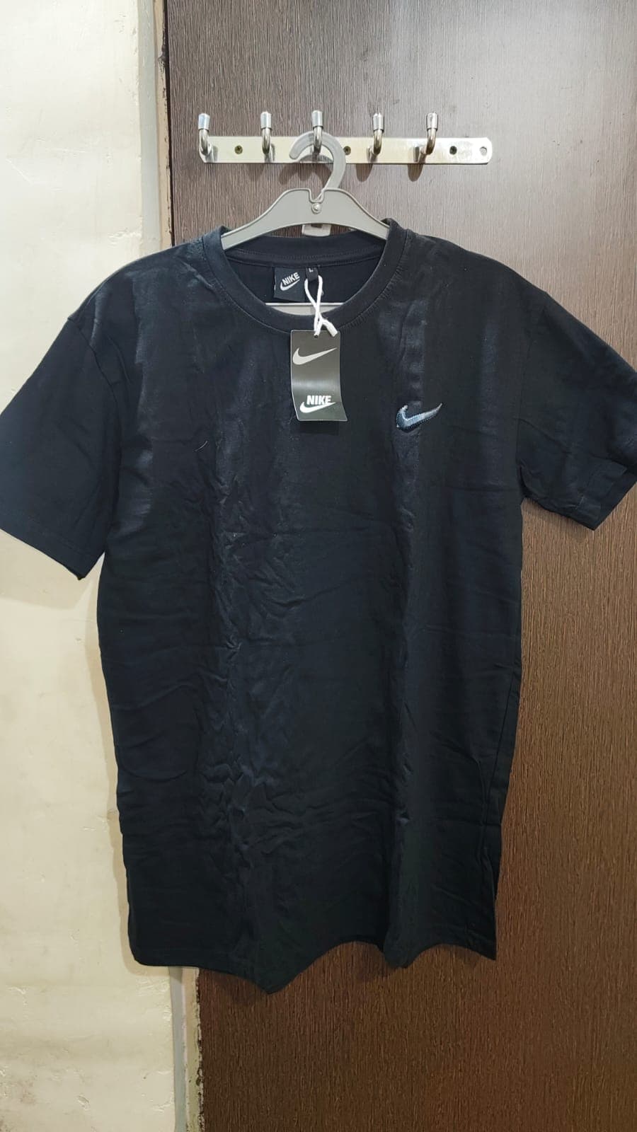 लकड़ी के दरवाजे पर हैंगर से लटकी हुई सिल्वर लोगो वाली काली नाइकी टी-शर्ट।