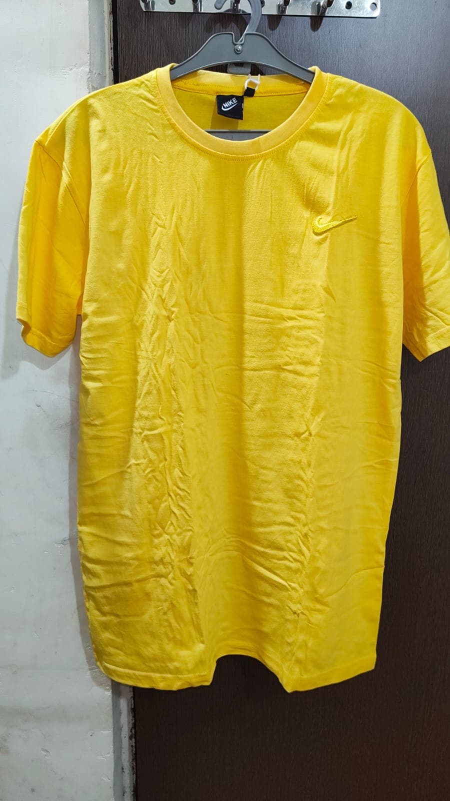 ग्रे हैंगर पर लटकी हुई चमकीली पीली नाइकी टी-शर्ट, जिस पर छोटा सा लोगो है।