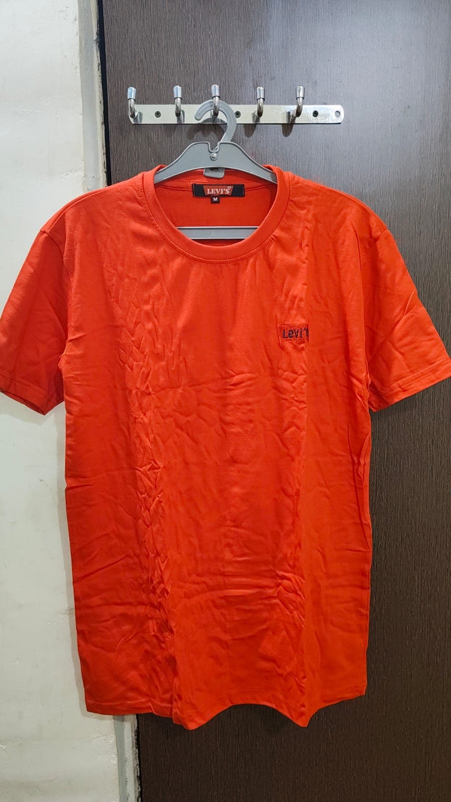 दरवाजे पर ग्रे हैंगर से लटकी हुई लेवाइस ब्रांड की चमकीली नारंगी टी-शर्ट।