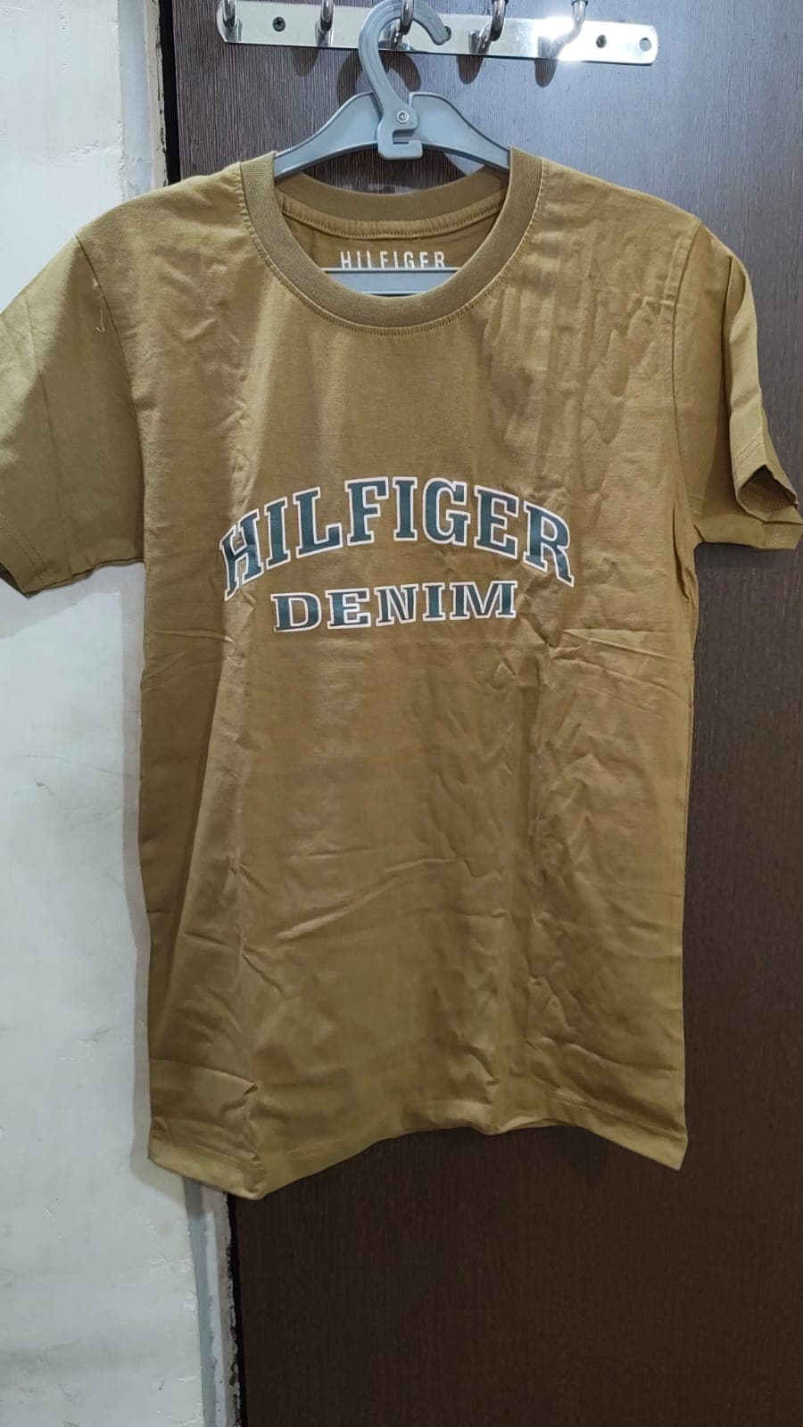हैंगर पर लटकी सरसों के पीले रंग की टी-शर्ट जिस पर 'Hilfiger Denim' लिखा है।